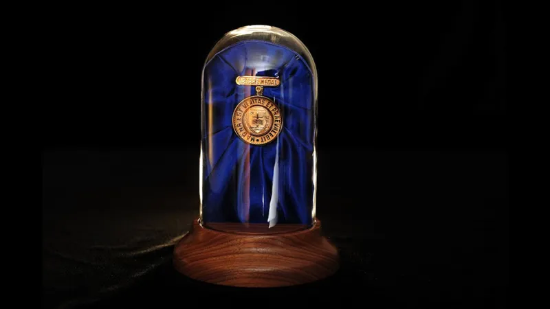 The gold Laetare Medal is displayed inside a clear glass dome on a wooden base. The medal is mounted against a dark blue fabric backdrop, and the entire display is set against a solid black background.